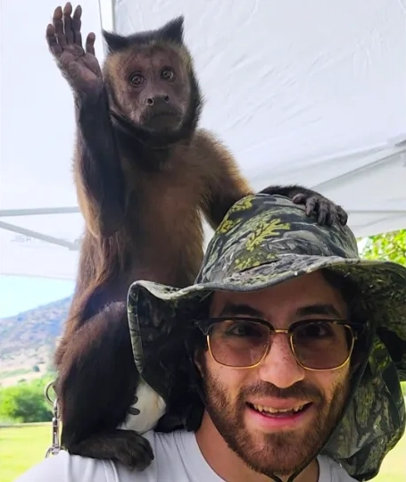 A friendly Zebra Entertainment animal handler smiles with a capuchin monkey waving from his shoulder during an event.