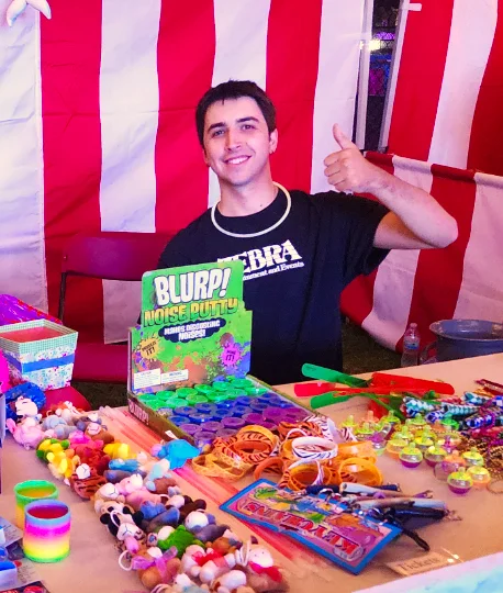 A smiling Zebra Entertainment staff member gives a thumbs-up behind a carnival booth counter overflowing with colorful game prizes.